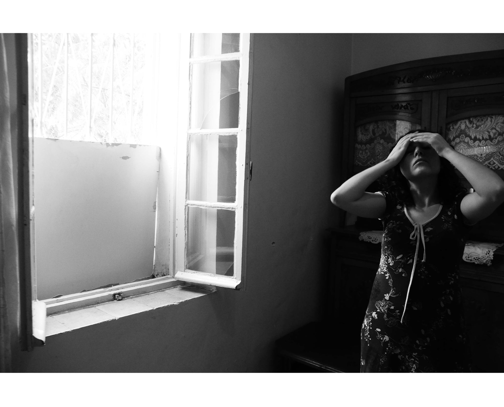 Une photo en noir et blanc d'une jeune femme nommée Lilia , les mains sur la tete devant une fenêtre entrouverte  